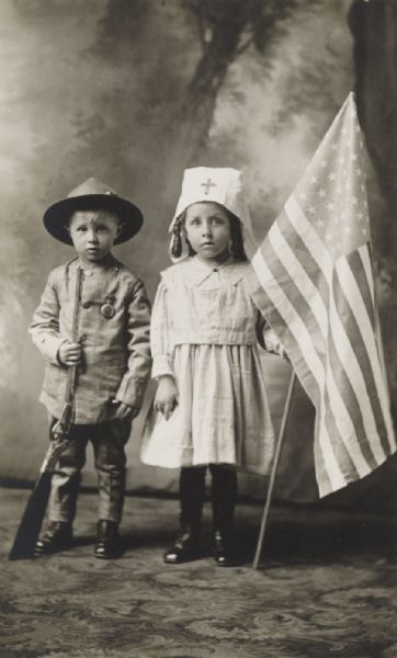 Full-length portrait of two children, about 6-7 years old, posed in a studio in front of a painted background. On the left the young boy is costumed as a soldier holding an air rifle, with a pocket watch pinned to the jacket. On the right the young girl is costumed as a Red Cross nurse holding a U.S. Flag.