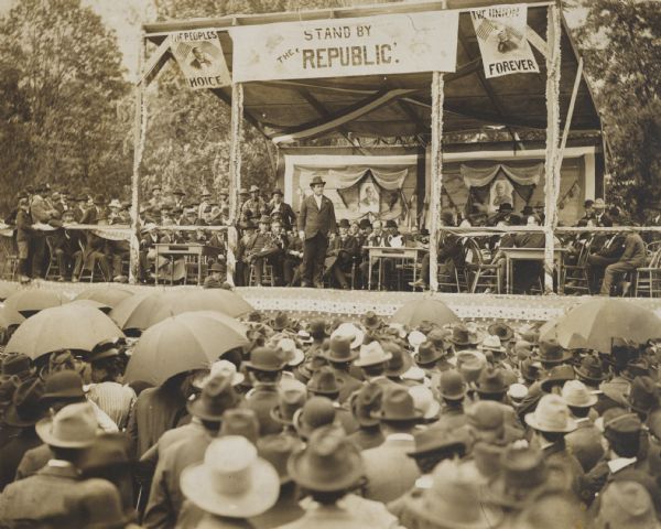 View from audience of William Jennings Bryan, as Democratic nominee for election to the presidency, addressing a crowd, estimated at 15,000 people, at a street fair. A large group of men are sitting and standing on the stage behind him. There are portraits of two men on a banner behind the stage.