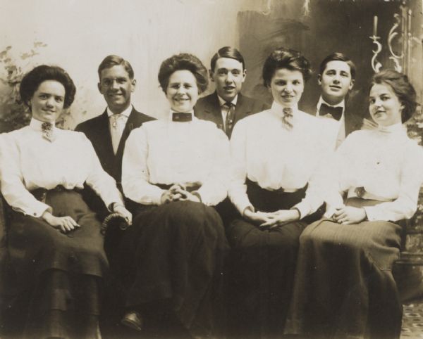 Group portrait of four women, seated in front of three men, all unidentified. The women wear high-collared white blouses and long dark skirts. The men wear suits and neckties. There appears to be a painted backdrop in the background.