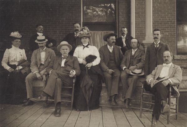 Group portrait of nine men and two women seated on a porch. A few of the men are holding cigars, and both of the women are holding umbrellas.