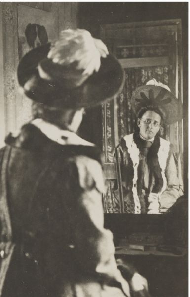 Unidentified woman posed sitting in front of a dressing table with a tall mirror. She is wearing eyeglasses, a hat with feathers, and a long coat with what appears to be a fur collar. On the wall on the left is a print of a dog.