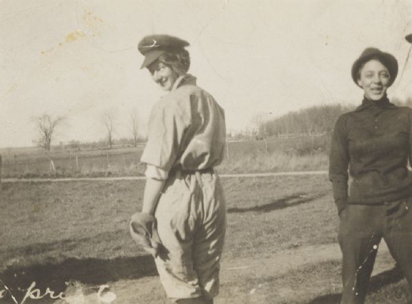 A girl standing in a field dressed in a man's baseball uniform. She is looking over her shoulder at the camera, and another woman wearing a hat and trousers stands beside her on the right.