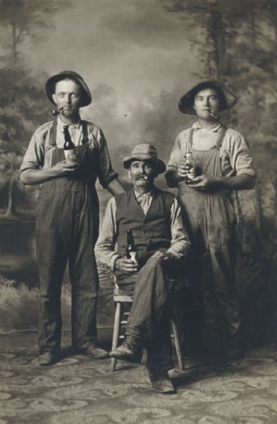 Studio portrait of three men smoking pipes and cigars and holding beer bottles in front of a painted backdrop. From left to right, Jacob Bowar, Herman Kalscheur, and Joseph Bowar.
