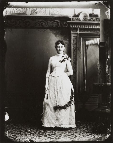 Full-length studio portrait in front of a painted backdrop of a woman posing standing, holding a hand fan, and wearing a corsage.