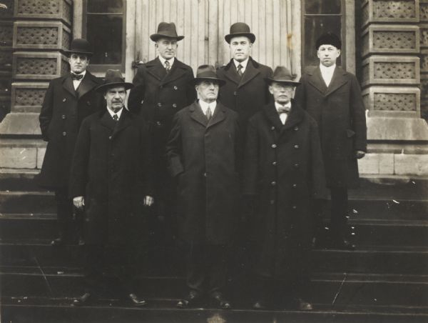 Posed group portrait of seven men standing on the steps of what appears to be a public building.
