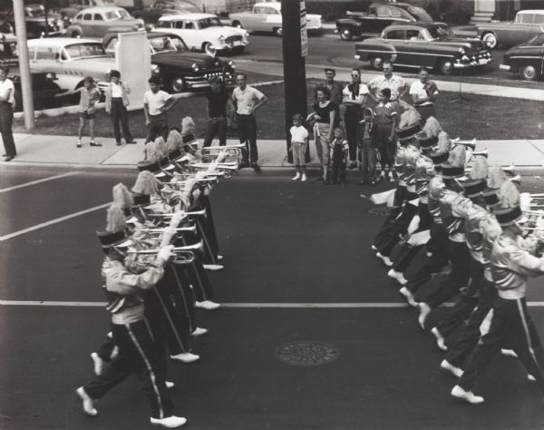 Uniformed boys' bugle corps passing along a street in a parade. A group of people watch from the sidewalk across the street.
