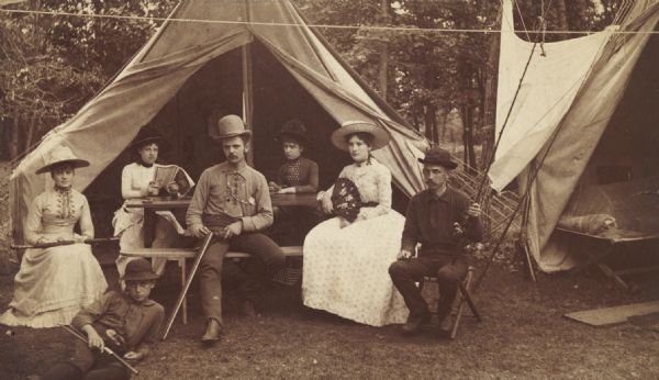 A group of young men and women posed before the tents of a hunting and fishing camp.