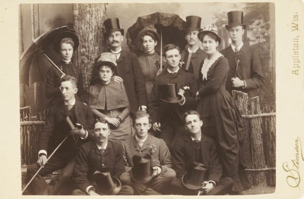 Posed group portrait of men and women members of the “Class of '86” at Lawrence College. The 8 men have silk top hats and the 4 women have umbrellas, for a semi-costumed effect.