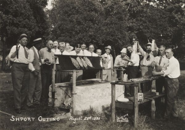 Gathering of men at a “Shport [sic] Outing”. The men are standing around a barbecue pit, preparing a feast of fish and corn.