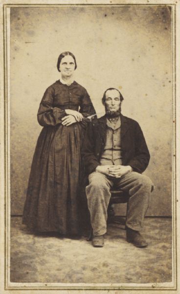 Carte-de-visite studio portrait of an unidentified couple. The man is seated, and the woman is standing beside him.