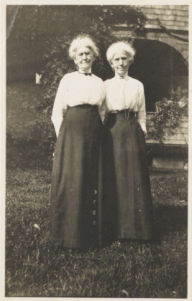 Portrait of Ellen Lloyd Jones and her sister Jane Lloyd Jones standing in front of the Hillside School which they founded and managed. Both sisters wear eyeglasses, high-collared white blouses, and long dark skirts. The Lloyd Jones sisters were aunts of the noted architect Frank Lloyd Wright.
