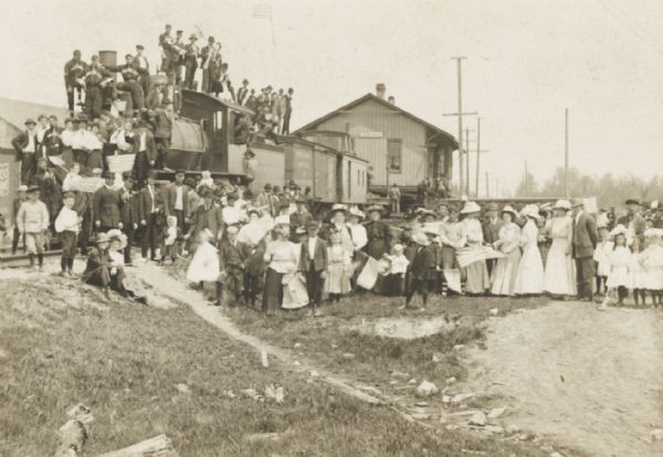 Gathering of people, some holding flags, celebrating the move of the small railroad depot along the track from Corinne to Gould City.

