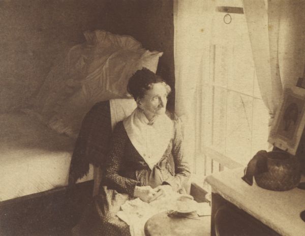 Mrs. Cadle seated in a bedroom, looking out a window. On the table in front of her is a cup and saucer.
