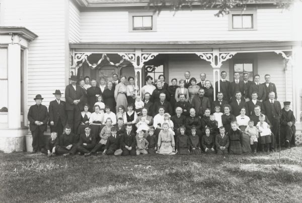 Outdoor group portrait of some eighty members of the extended Meinen family on the occasion of the golden anniversary of two of its members. They are standing on and around the porch of a house.