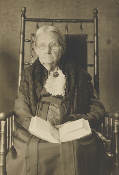 Portrait of elderly woman sitting in a chair, holding a book in her lap.