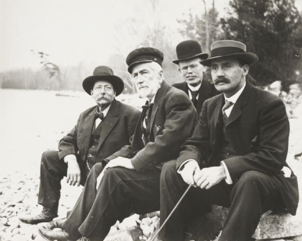 Four men seated on rocky shoreline of a lake in front of D.S. Crandall's house. Left to right: E.A. Corneille, D.S. Crandall, Harry E. Dankoler, and Joe Leighton. Harry Dankoler holds the shutter release.