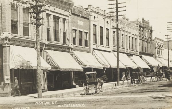 Photographic postcard of a view along left side of Monroe Street, showing horse-drawn vehicles parked along the curb in front of storefronts. Caption reads: "Monroe St., Waterloo, Wis."
