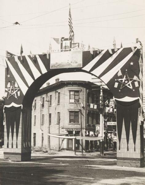 Decorated arch erected on State Street as part of the Semi-Centennial celebration of statehood. The arch bears a representation of Commodore Dewey's victory at Manila Bay in 1898. Signs on the brick building in the background read: "State Saloon," and "Labor Hall."