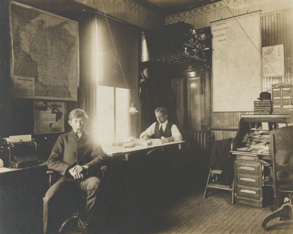 Interior, with two men, of a small office, very likely a railroad company office. Maps are on the wall, and rolled up on a shelf near the ceiling.