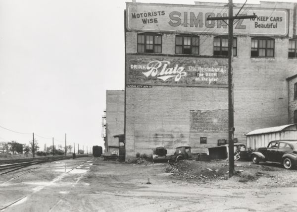 View down side of railroad tracks towards commercial buildings. Two advertisements are painted on the side of the large brick building in the foreground.