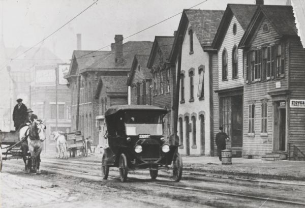 Street view of the north side of State Street, west of 6th Street. The second frame house from the right was once occupied as an office by Victor L. Berger (former Mayor of Milwaukee, famous Socialist). The Ford touring automobile in the foreground (according to annotation) was “one of the first jitney busses”. Independent owners put a sign in the windshield showing destination and charged random passengers for a ride there.