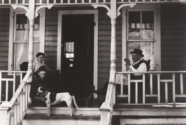 Family on the front porch of a small house, relaxing, reading, with dog. The photograph was probably made as an illustration for the University of Wisconsin College of Agriculture.

