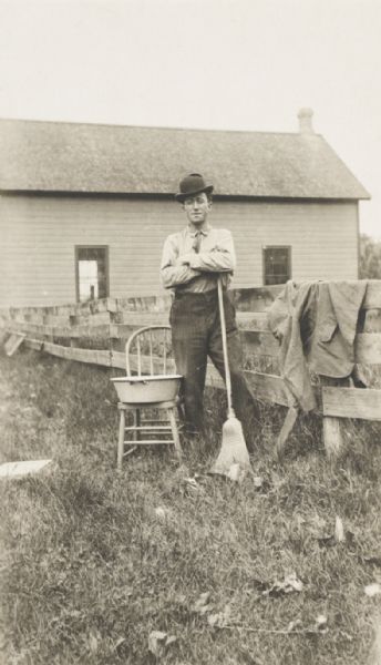Unidentified man standing in (a farmyard?) with a broom and wash-basin. 
