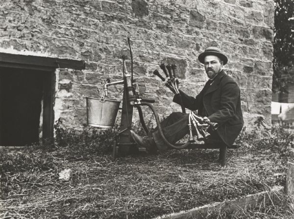 Unidentified man (inventor?) seated at and showing a foot-powered (?) milking machine. He is outdoors near a stone building.