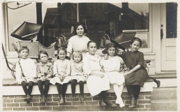 Seven children and one young adult, posed, seated in front of a (store?) window. Behind the group are baby carriages. One child is hiddenbehind the girl on the right.