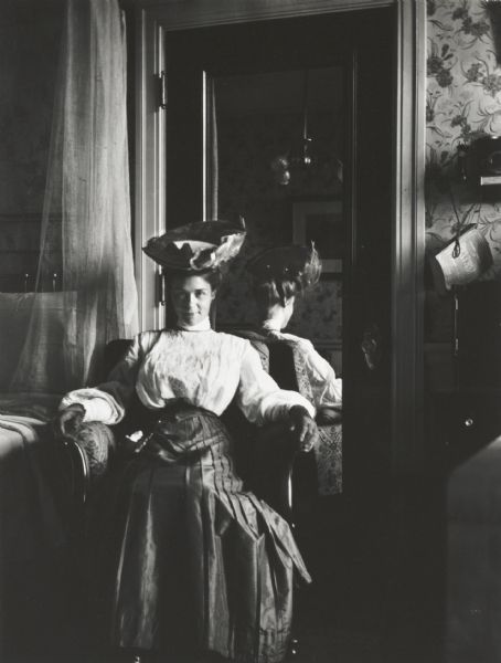 Mary E. Smith seated in a bedroom in the Belvedere Hotel (?), probably in Washington, D.C. or vicinity. Her reflection is in the mirrored door behind her. To the right of the door is a telephone with a Chesapeake & Potomac telephone directory.