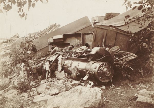 View looking down at men gathered at railroad wreck The locomotive is on its side, and the freight cars are demolished.