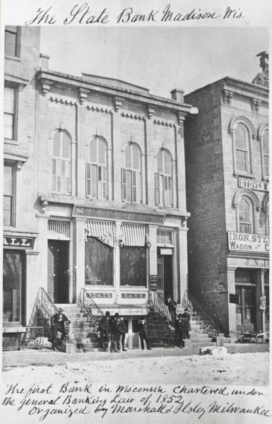 External view of the State Bank, the first bank in Madison. A group of men are posed along the sidewalk in front.
