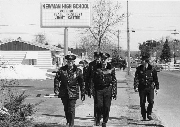 President Jimmy Carter's visit meant extra duty for the local police force. An elevated sign in the background reads "Newman High School, Welcome, Peace President, Jimmy Carter." There are barricades and cars in the streets and trees and homes are in the background. Snow is on the ground.