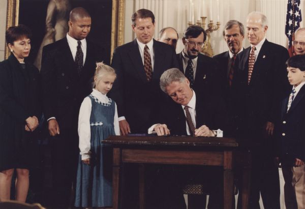 President Bill Clinton signing the FY 1998 Labor, HHS, and Education Appropriation Bill. Behind the President are Vice President Al Gore and Wisconsin Congressman David R. Obey, chair of the House Appropriations Committee. Secretary of Labor Alexis Herman is standing at the far left.