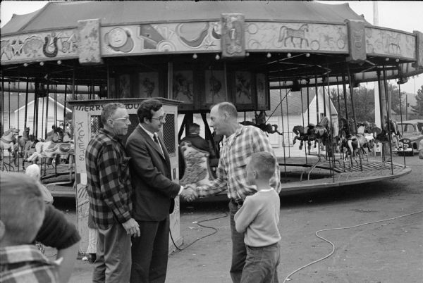Running for re-election for the first time, Congressman David R. Obey greets voters at the fair. A merry-go-round is in the background. Although unidentified, it is likely this is the Forest County Fair.