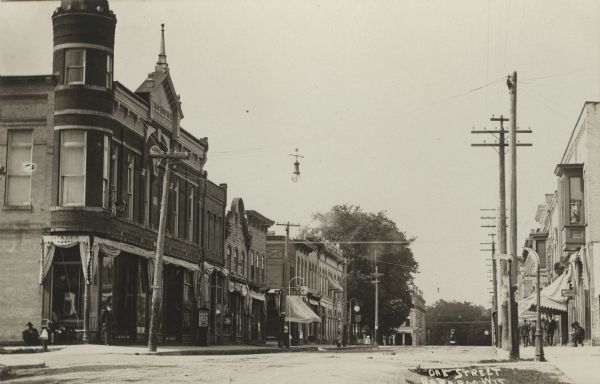 Photographic postcard view from street in the central business district. The street is lined with businesses, and a few have awnings over their storefronts. Pedestrians are on the sidewalk on the right. Caption reads: "Oak Street, Baraboo, Wis."