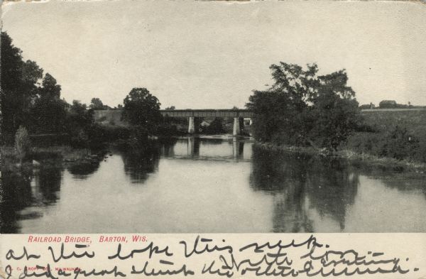 View over water towards a railroad bridge over a river. Trees and shrubs are along the banks of the river. There are buildings in the distance behind the bridge. Caption reads: "Railroad Bridge, Barton, Wis."