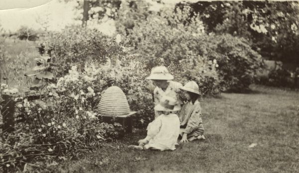 Miss Dorothy Quincy Wright and two children sit on the ground next to a bee skep in yard surrounded by flowers. Linden apiary.