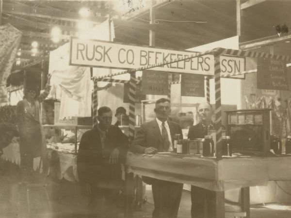 Group of men at the Rusk County Beekeepers Association, standing near table of honey display. Center - R. Knudtson, Glen Flora, President. Right - A. D. Calkins, Ladysmith, Secretary-Treasurer. Left - Leslie Yancey, Ladysmith, Vice-President.