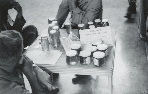Three men in conversation around several jars of honey on a table. Sign reads: "Wisconsin Honey Market Day." Written on back: "A typical scene of one of the producer's tables on display."