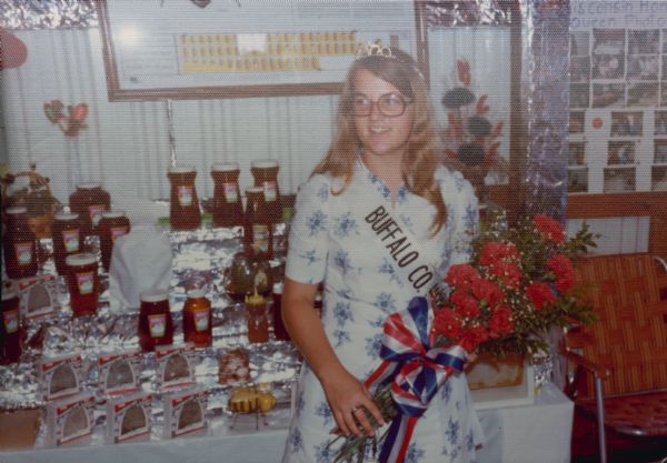 LeAnn Wenger, the Buffalo County Honey Queen. Wenger's hometown is Fountain City, Wisconsin. She is standing in front of a honey display, wearing a Buffalo County sash, holding a bouquet, and wearing a tiara.