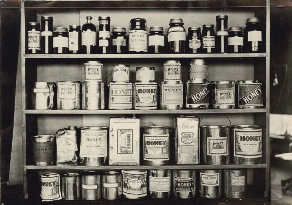 Display of honey containers with printed labels on four wooden shelves. On the top shelf are glass jars of honey, and the bottom three shelves have a variety of cans and tins.