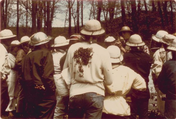 View from behind of a man with a small swarm of bees attached to the back of his sweater. He is standing at the back of an outdoor beekeeping class. About a dozen people are in the class and many are wearing bee hats with veils.