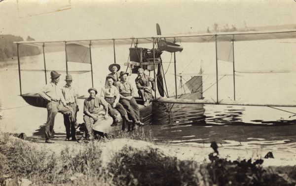View from shoreline of Trout Lake of a group of six men posed with the pilot, possibly Jack Vilas, on an early seaplane or flying boat floating in the water at the shoreline. The six men are dressed in work clothes and hats.<p>This is an early use of an aircraft to detect forest fires.