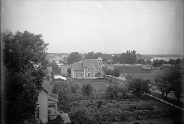 Elevated view of houses and yards on Main Street between 2nd and 3rd Avenues. There is a large white house with a tower is the family home of the Walters who own the "pop" factory. A brick structure, the Scott Store, is across Main Street, one of the first in Wisconsin. Trees line the street and mark the boundaries of the Walters' yard. In the background is a bridge over a lake.
