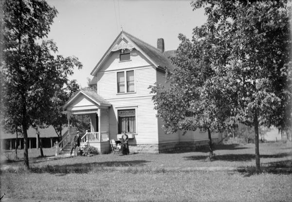 View across front lawn of family posed in yard of house located on S. 3rd Street. Otto Ansorge leans on the bannister of his front porch. An unidentified woman (probably his wife) stands in front of the house's front window besides a baby sitting in a stroller. There are trees in the yard, and a board sidewalk in front of the house. Behind the house on the left is a shed or garage.