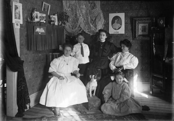 The Bardes Family poses in their home. Family members pictured include (left to right): Nora, Jerry, Alice, Mrs. Bardes, and Barbara. The portrait also includes the family dog sitting in the middle on a footstool. The room is decorated with wallpaper, lace, and various portraits. The home belonged to Don Bardes (not pictured), a barber.