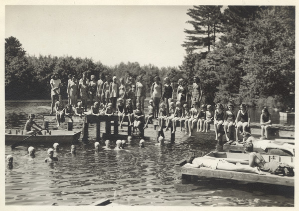 Water scene, with many campers in swimsuits lined up on a dock, some standing, some sitting. A number of campers are also in the water, wearing swim caps. One camper is sitting in a rowboat, and other campers are sunbathing on two smaller docks. Original caption notes: "Scene at the waterfront in 1943 during a lull in the swimming instruction period."
