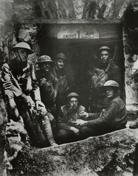 Allied soldiers gathered near an entrance to an unknown establishment. A sign in French above the entrance is not legible, but there is an image of a cricket and the words: "Le Grillon." The man squatting in the middle is smoking a pipe, and the man next to him appears to be holding a grenade in each of his hands.