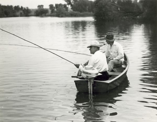 Two men fishing from a small boat powered by an Evinrude outboard motor. One of the men is smoking a pipe. In the background is a wooded shoreline.